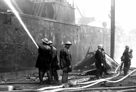 Surrey Commercial Docks, Blitz WW2. A burnt-out cargo ship in the Surrey Commercial Docks, Rotherhithe.  Firefighters continue to damp down hot-spots of fire within the ship.  X..png